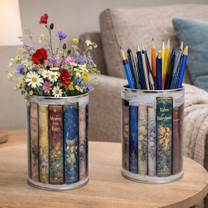 May include: Two cylindrical containers with a distressed white finish and a book-themed design. One holds colorful wildflowers, the other pencils and pens. The containers are on a wooden table, with a lamp and sofa in the background.