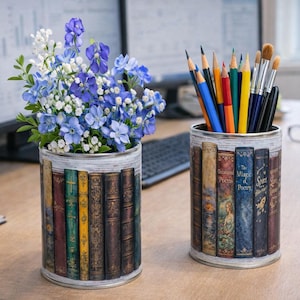 May include: Two decorative tin cans, one filled with blue and white flowers, the other with pencils and brushes. The cans are wrapped with a design of antique books. The cans sit on a wooden desk.