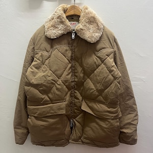 May include: A tan quilted jacket featuring a beige faux fur collar, a full-length front zipper closure, and two large rectangular front pockets. The garment has a diamond-stitched pattern across the chest and long sleeves with simple cuffs.