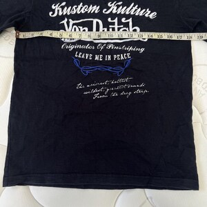 May include: Black t-shirt with white and blue text. The text includes "Kustom Kulture Von Dutch" and "Originator Of Pinstriping LEAVE ME IN PEACE". Additional script is also visible. The shirt is being measured with a tape measure.