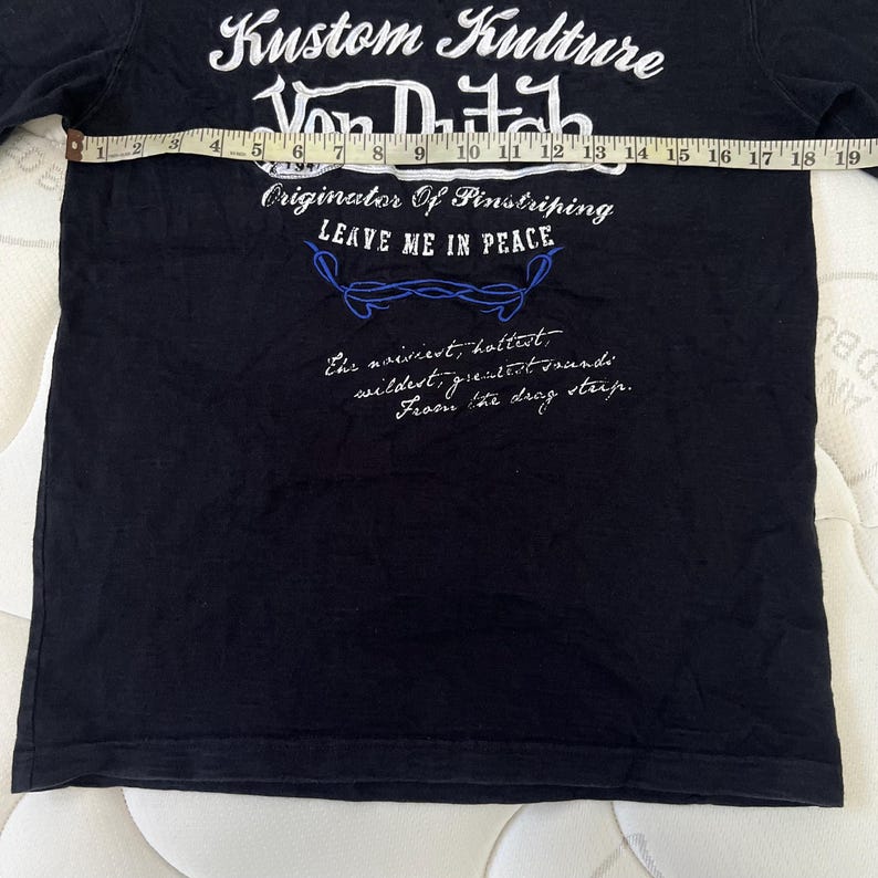 May include: Black t-shirt with white and blue text. The text includes "Kustom Kulture Von Dutch" and "Originator Of Pinstriping LEAVE ME IN PEACE". Additional script is also visible. The shirt is being measured with a tape measure.