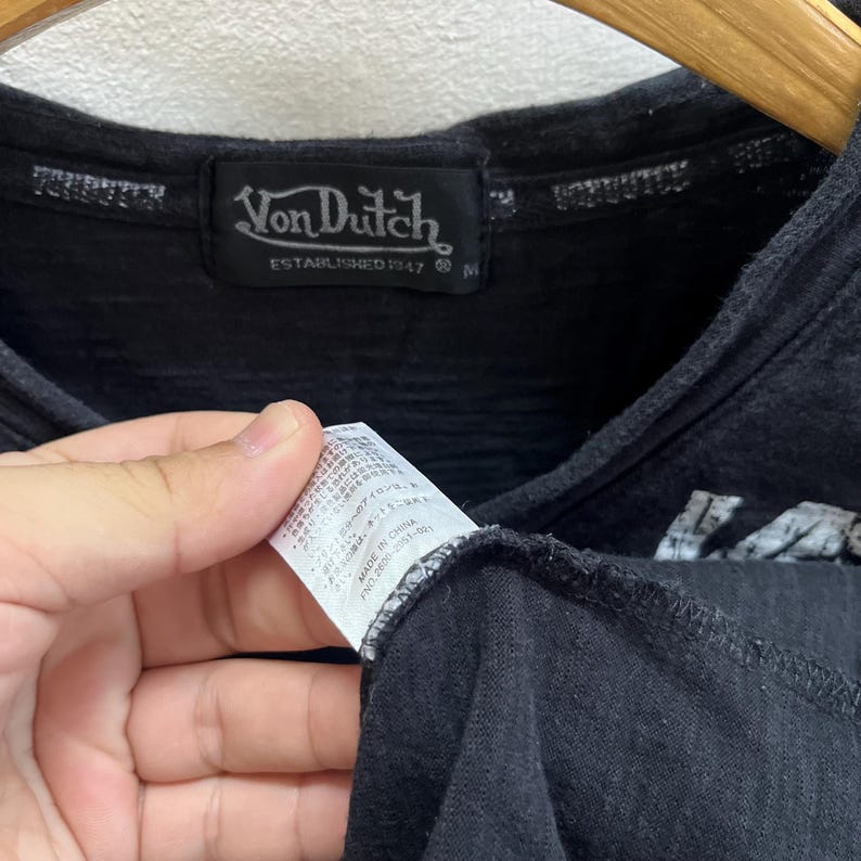 May include: Black V-neck shirt with the Von Dutch logo on a black label. The label reads "Von Dutch ESTABLISHED 1947". A tag with Japanese text is held in the frame.