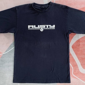 May include: Navy blue t-shirt with the word "RUSTY" in white block letters across the chest. Below the brand name are the words "FOUNDED ON INNOVATION" and "FOCUSED ON THE FUTURE" in white. The shirt has short sleeves and a crew neck.