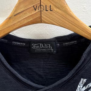 May include: Black V-neck shirt with a Von Dutch label. The label is black with white text and the words "Von Dutch ESTABLISHED 1947". The shirt is hanging on a wooden hanger.