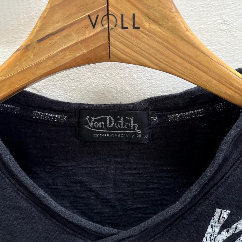 May include: Black V-neck shirt with a Von Dutch label. The label is black with white text and the words "Von Dutch ESTABLISHED 1947". The shirt is hanging on a wooden hanger.