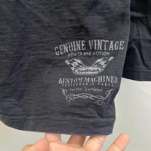 May include: Close-up of a black t-shirt featuring a vintage graphic. The graphic includes the text "GENUINE VINTAGE POWER and MOTION", a checkered flag design, and "Von Dutch" branding. Below, it reads "KUSTOM MACHINED PERFORMANCE PARTS Est 1947 Compton".