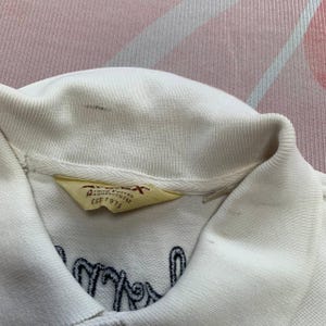 以下が含まれることがあります： 襟と刺繍テキストが付いたクリーム色のポロシャツ。シャツには「Armed Forces Manufacturer EST. 1975」と書かれたタグが付いています。刺繍されたテキストは濃い青色です。