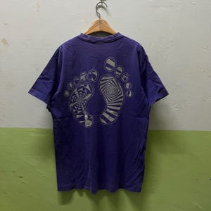 May include: A purple t-shirt featuring a graphic of two feet on the back, each with a unique design. One foot has a spiral pattern, the other a geometric design. The shirt hangs on a wooden hanger.