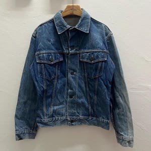 Può includere: Una giacca in denim blu a lavaggio medio con colletto a punta, chiusura frontale con bottoni e due tasche sul petto con patta abbottonata. La giacca presenta maniche lunghe con polsini abbottonati e cuciture verticali sul davanti.