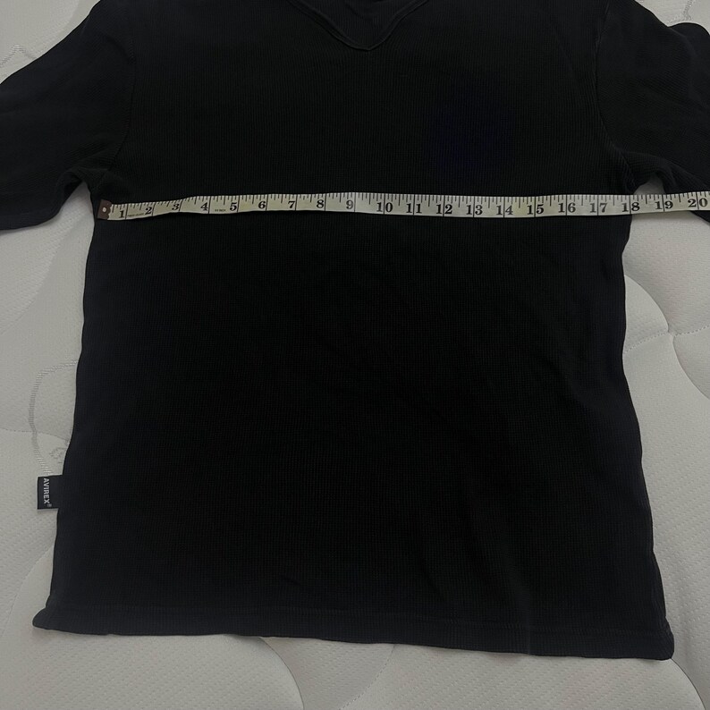 May include: A black short-sleeved shirt with a ribbed texture and a V-neckline. A white measuring tape is placed horizontally across the chest, showing a width measurement of approximately 51 cm from edge to edge. A small black tag reads Avirex.