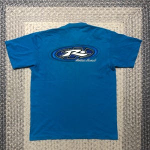 May include: A turquoise blue t-shirt with a graphic on the back. The graphic is an oval shape with a blue and white design and the letters "R" and "S" inside. Below the oval is the text "Custom Brand" in a cursive font.