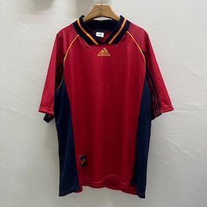May include: Red and navy blue Adidas soccer jersey with yellow accents. The jersey has a V-neck collar and the Adidas logo in gold. The sleeves are short and feature navy blue stripes. The jersey has a tag on the lower left side.
