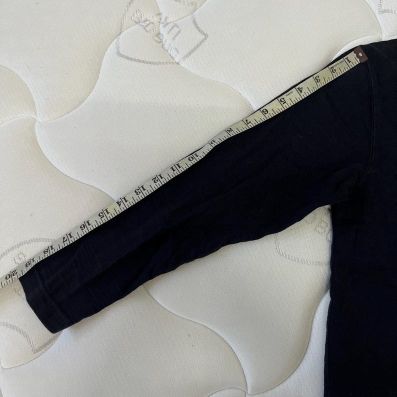 May include: A black long-sleeved shirt with a measuring tape across the sleeve. The tape shows measurements up to 50 cm. The shirt is on a white quilted surface.
