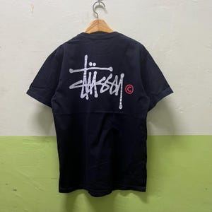 May include: Black short-sleeved t-shirt featuring a white logo and a red copyright symbol. The logo is a stylized signature. The t-shirt is suspended on a wooden hanger against a green and yellow backdrop.