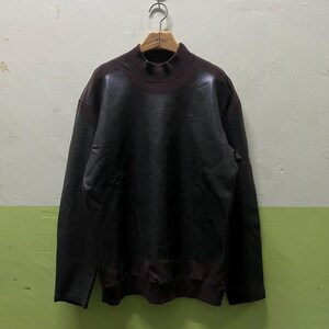 May include: A dark brown mock neck sweatshirt with a leather-like appearance. The long-sleeved top is hanging on a wooden hanger. The sweatshirt has a ribbed collar and cuffs. The background is a light green and beige wall.