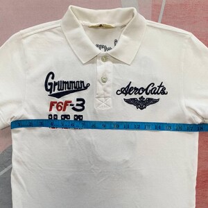 以下が含まれることがあります： 襟付きで半袖の白いポロシャツ。「Grumman」や「Aero Cats」などの刺繍テキストとグラフィックが特徴です。 シャツの上にメジャーが置かれており、約46cmの測定値を示しています。