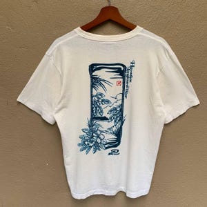 May include: White t-shirt with a blue rectangular design on the back, depicting tropical plants, a bird, and the text "Hawaiian Longboard Wear." The shirt has short sleeves and a crew neck.