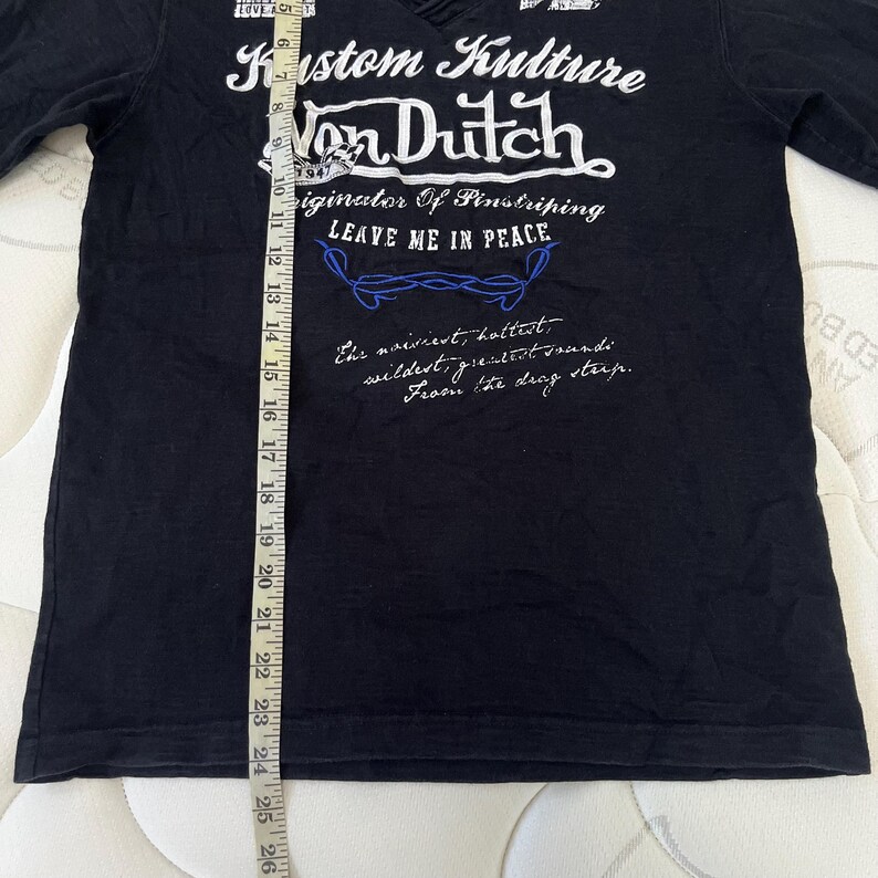 May include: Black V-neck t-shirt with white text that reads "Kustom Kulture Von Dutch" and "LEAVE ME IN PEACE". The shirt has a blue barbed wire design and additional text. The shirt measures approximately 63.5 cm in length.