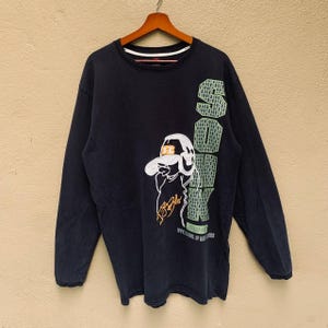 Könnte beinhalten: Marineblaues Langarmshirt mit einer Cartoon-Figur, die einen Hut trägt, und dem Wort "SOOK" in grünen Blockbuchstaben. Das Shirt hat auch den Text "www.school of hawks".