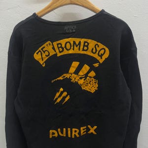 May include: A black long-sleeve thermal shirt featuring a yellow graphic on the back. The graphic includes the text 75TH BOMB SQ above an illustration of a stylised aircraft and lightning bolts. The word AVIREX is printed in yellow at the bottom.