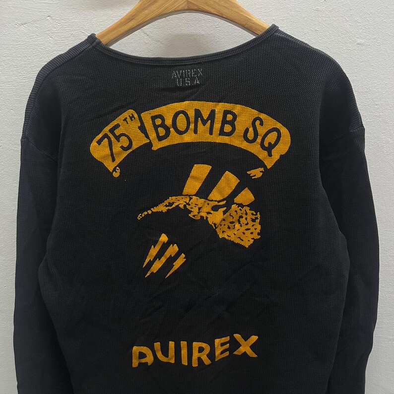 May include: A black long-sleeve thermal shirt featuring a yellow graphic on the back. The graphic includes the text 75TH BOMB SQ above an illustration of a stylised aircraft and lightning bolts. The word AVIREX is printed in yellow at the bottom.