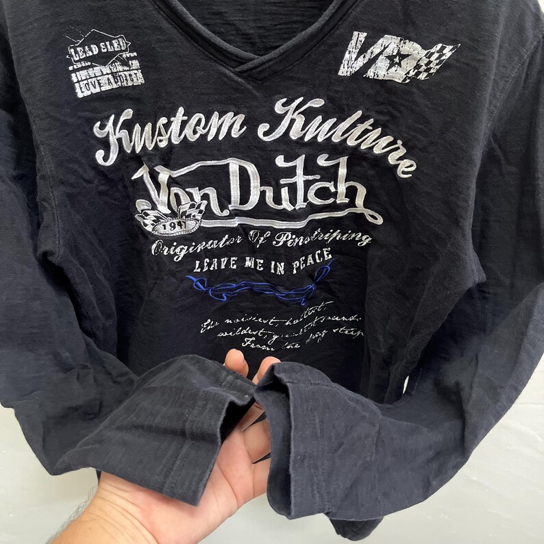 May include: Black long-sleeved V-neck shirt featuring the text "Kustom Kulture Von Dutch" in white, along with other graphics and text. The shirt also displays the phrases "LEAVE ME IN PEACE" and "Originator Of Pinstriping".