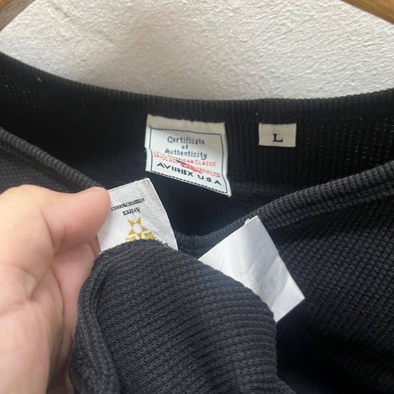 May include: A black thermal long-sleeve shirt with a waffle-knit texture. The interior collar features a white tag labelled Certificate of Authenticity Avirex U.S.A. and a separate white tag with the size L. An additional white tag shows the Avirex logo.