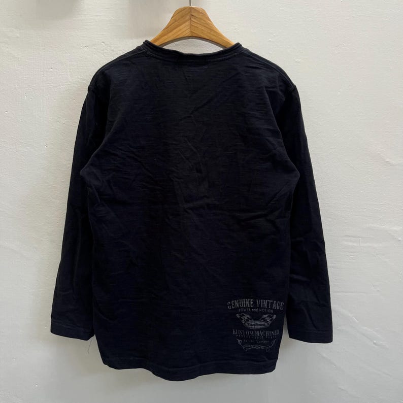 May include: Black long-sleeved top with a crew neck. The shirt has a faded graphic on the lower back that reads "GENUINE VINTAGE" and "CUSTOM MACHINES". The top is hanging on a wooden hanger.