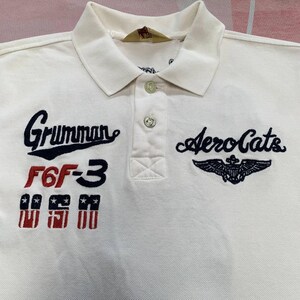 以下が含まれることがあります： 襟付きでボタンが3つ付いたクリーム色のポロシャツ。「Grumman F6F-3」と翼のエンブレムが付いた「AeroCats」の刺繍入り。クラシックでカジュアルなスタイルです。