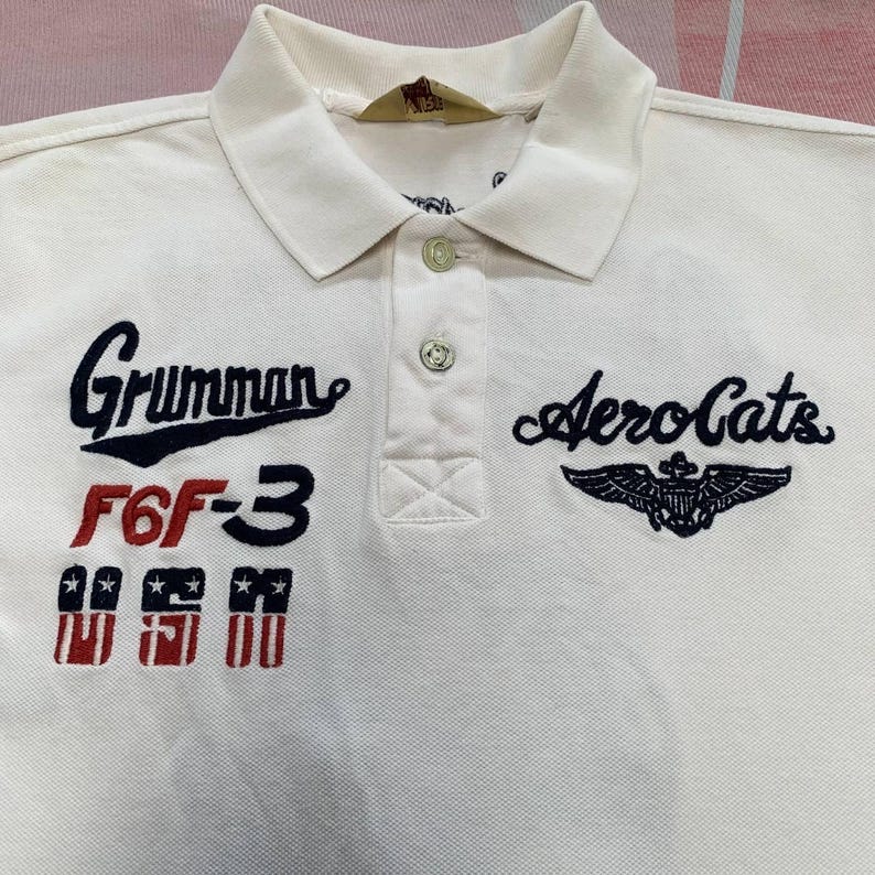 以下が含まれることがあります： 襟付きでボタンが3つ付いたクリーム色のポロシャツ。「Grumman F6F-3」と翼のエンブレムが付いた「AeroCats」の刺繍入り。クラシックでカジュアルなスタイルです。