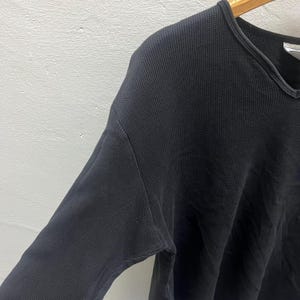 May include: A long-sleeved shirt in a dark charcoal grey colour featuring a textured waffle-knit fabric. The garment has a rounded neckline with a ribbed trim and is displayed on a wooden hanger, showing the shoulder and sleeve construction.