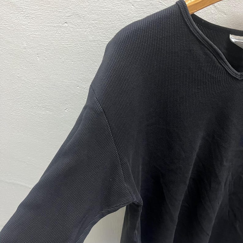 May include: A long-sleeved shirt in a dark charcoal grey colour featuring a textured waffle-knit fabric. The garment has a rounded neckline with a ribbed trim and is displayed on a wooden hanger, showing the shoulder and sleeve construction.