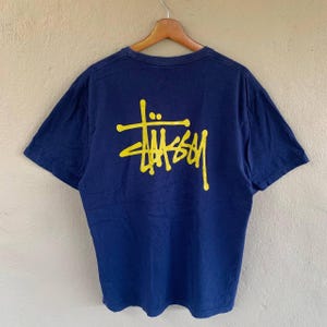 May include: A navy blue t-shirt with a yellow logo on the back. The logo is a stylized script design. The shirt is hanging on a wooden hanger. The t-shirt is made of a soft material.