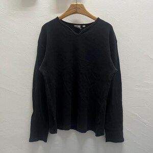 May include: A solid black long-sleeve shirt featuring a V-neckline with a ribbed trim detail. The garment is constructed from a textured fabric and is displayed on a wooden hanger, showcasing a simple design with long sleeves and a straight hem.