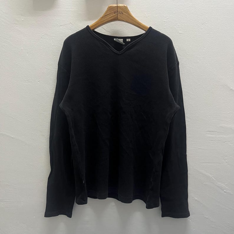 May include: A solid black long-sleeve shirt featuring a V-neckline with a ribbed trim detail. The garment is constructed from a textured fabric and is displayed on a wooden hanger, showcasing a simple design with long sleeves and a straight hem.