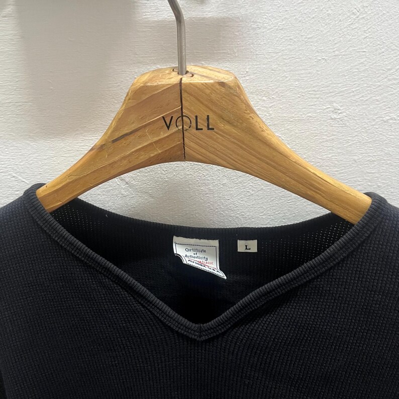 May include: A black V-neck shirt with a textured waffle-knit fabric hanging on a wooden hanger labelled VOLL. The interior collar features a white tag with the text Certificate of Authenticity and a separate white tag with the letter L.