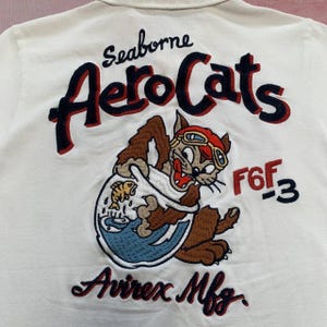 以下が含まれることがあります： 白いポロシャツ。刺繍された「Seaborne AeroCats」のデザイン。飛行士ゴーグルをかけた漫画の猫が金魚鉢を持っており、「F6F-3」と「Avirex Mfg.」の文字があります。赤、ネイビー、茶色、金色の糸を使用しています。