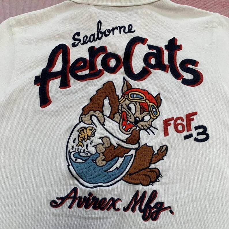 以下が含まれることがあります： 白いポロシャツ。刺繍された「Seaborne AeroCats」のデザイン。飛行士ゴーグルをかけた漫画の猫が金魚鉢を持っており、「F6F-3」と「Avirex Mfg.」の文字があります。赤、ネイビー、茶色、金色の糸を使用しています。