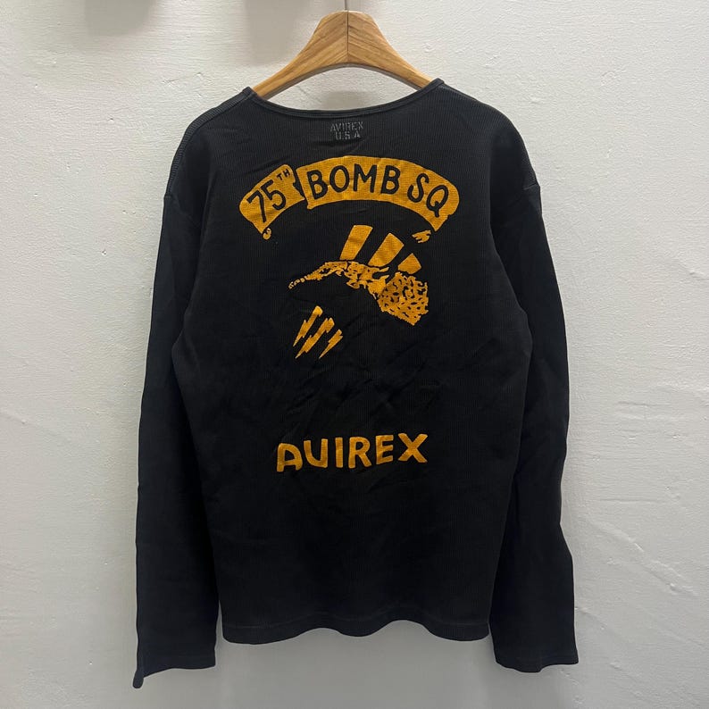 May include: A black long-sleeve shirt featuring a yellow graphic on the back. The graphic includes the text 75TH BOMB SQ, an illustration of a bird, and the word AVIREX. The shirt has a crew neckline and is displayed on a wooden hanger.
