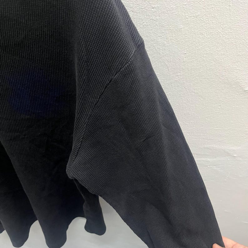 May include: A long-sleeved shirt made of a black waffle-knit fabric. The garment features a textured grid pattern throughout the material, a crew neckline, and a relaxed fit with dropped shoulder seams that extend down the arm to the sleeve.