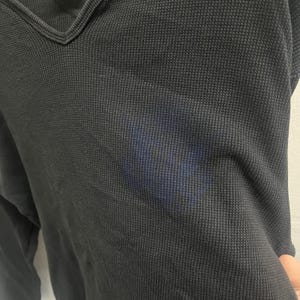 May include: A black long-sleeve shirt with a V-neckline and a textured waffle-knit fabric pattern. The garment features a subtle, darker blue rectangular discoloration or stain visible on the front chest area of the fabric.