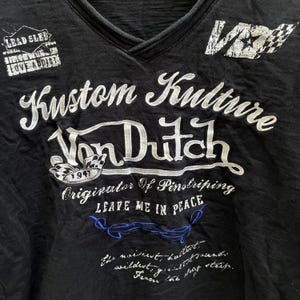 May include: Black V-neck t-shirt with white embroidered text reading "Kustom Kulture Von Dutch" and other phrases. The shirt features patches saying "Lead Sled" and "Love Addicts". A checkered flag graphic is also visible.
