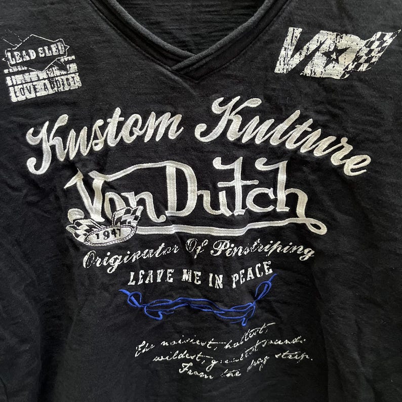 May include: Black V-neck t-shirt with white embroidered text reading "Kustom Kulture Von Dutch" and other phrases. The shirt features patches saying "Lead Sled" and "Love Addicts". A checkered flag graphic is also visible.