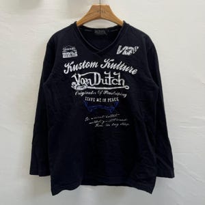 May include: Black long-sleeved V-neck shirt featuring white graphic text including "Kustom Kulture Von Dutch". The shirt has a vintage design and is made of a soft fabric. The text also includes the phrase "Leave me in peace".