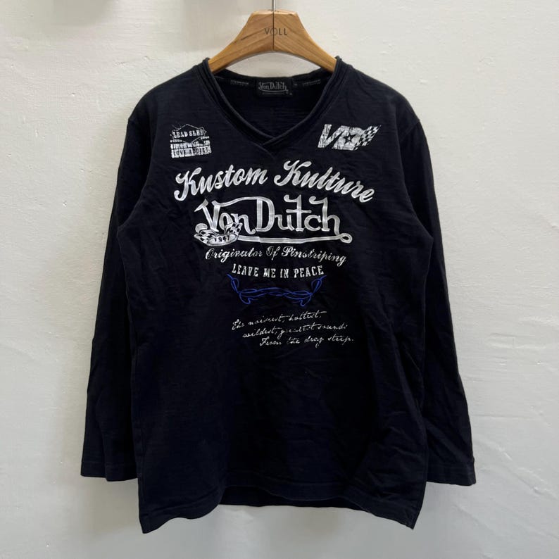 May include: Black long-sleeved V-neck shirt featuring white graphic text including "Kustom Kulture Von Dutch". The shirt has a vintage design and is made of a soft fabric. The text also includes the phrase "Leave me in peace".
