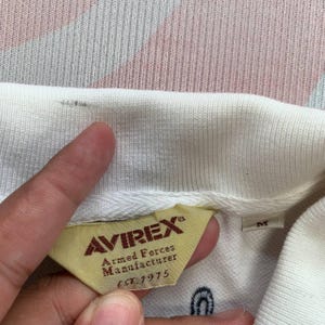 以下が含まれることがあります： ブランドタグ「AVIREX Armed Forces Manufacturer EST. 1975」が付いた白いポロシャツの襟のクローズアップ。 タグはタン色で、赤と黒の文字が書かれています。 小さな「M」タグが見えます。