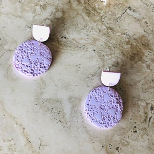 May include: A pair of lavender-colored earrings with a circular design. The earrings have a white backing and a delicate, intricate pattern.