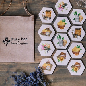 May include: A "Busy Bee Memory Game" set with hexagonal wooden tiles featuring bee-related illustrations. A canvas bag with the game's title and a bee graphic is on the left. A bundle of lavender is at the bottom.