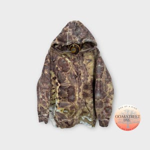 Könnte beinhalten: Ein brauner und grüner Tie-Dye-Hoodie mit einer Kapuze mit Kordelzug. Der Hoodie hat ein Etikett mit der Aufschrift "OOAKSTREET DYE" und "ONE OF A KIND" in einem Kreis.