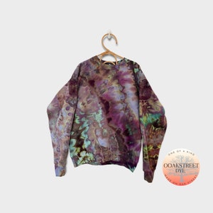 Puede incluir: Una sudadera tie-dye en tonos de morado, marrón, verde y azul. La sudadera tiene cuello redondo y mangas largas. Está colgada de una percha de madera. La etiqueta de la sudadera dice "Oak Street Dye, One of a Kind, Made on Oak Street."